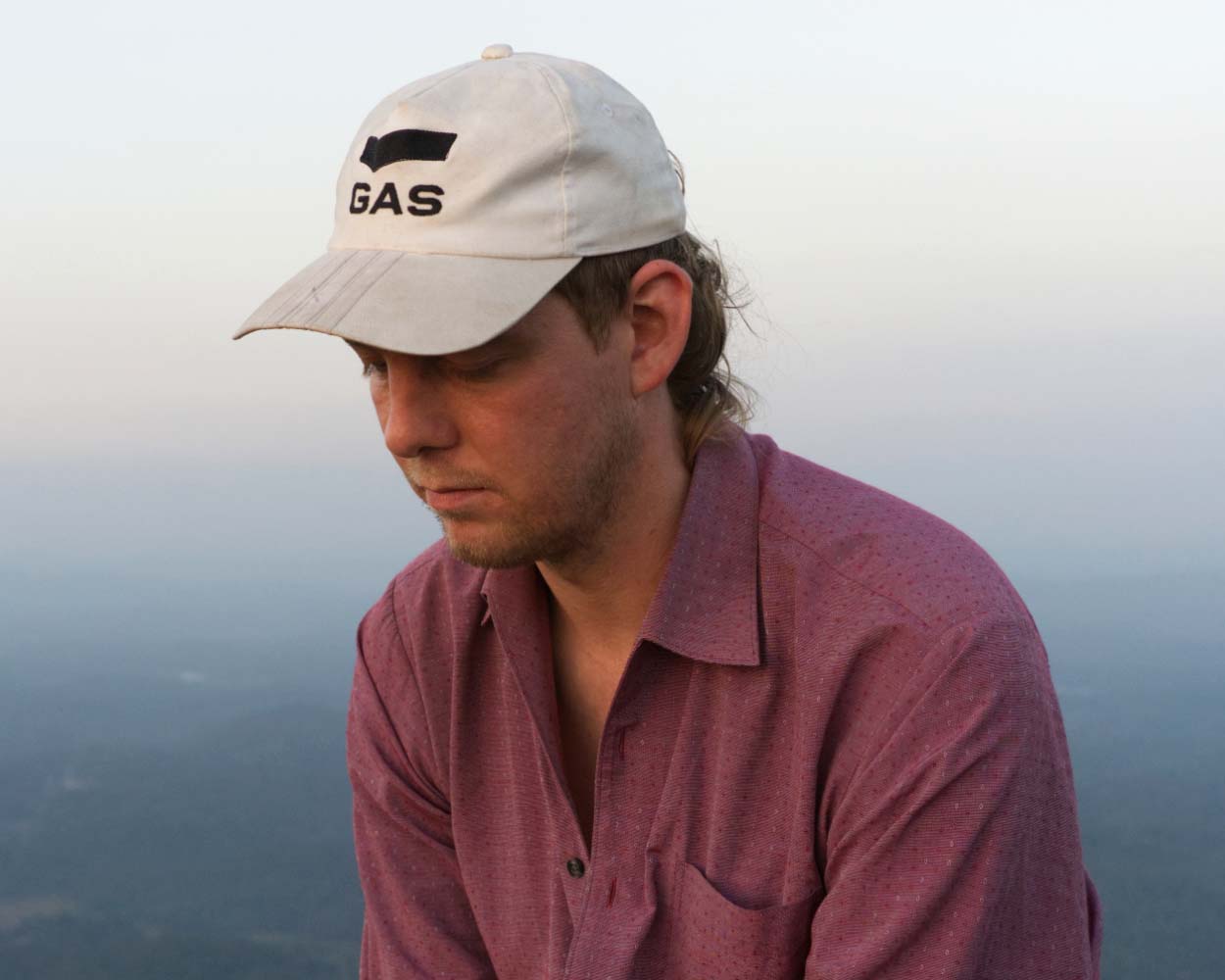Head and shoulders of Felix Deufel, solemnly looking downwards, wearing a white baseball cap, against a distant background of haze and mist.