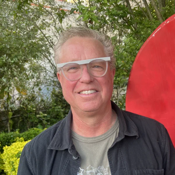 Square portrait of a smiling person with light skin, grey hair, dark-rimmed glasses, and a gray T-shirt in front of greenery and a bright red object on the right.