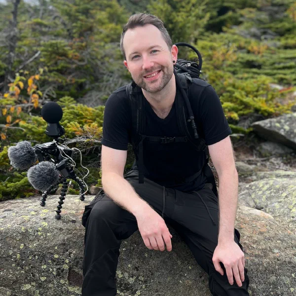 Portrait of a smiling light-skinned person seated on a lichen-covered boulder next to a field recording rig with evergreens behind.