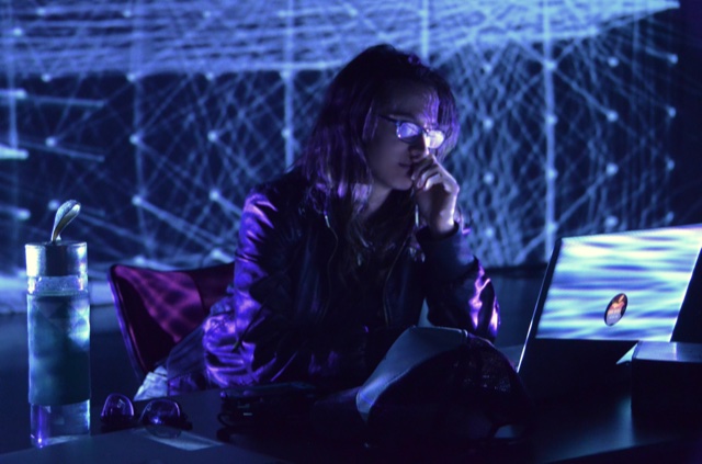 Monica, a young woman with glasses, is sitting at a computer with network node projections behind her