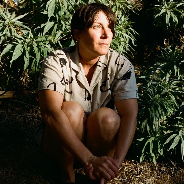 Light-skinned person crouched among green plants in dappled sunlight; she is wearing a taupe-colored shirt with black brushstrokes printed on it.